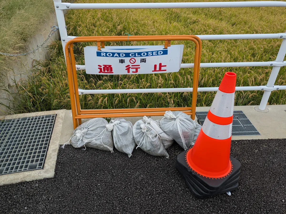 道路の脇に「ROAD CLOSED 車両通行止」の柵と、土のう、コーンが置かれている